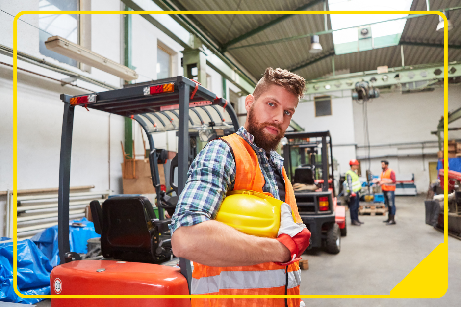 In die Kamera schauender Mann mit Sicherheitsweste und Helm in einer Lagerhalle vor einem Gabelstapler stehen.